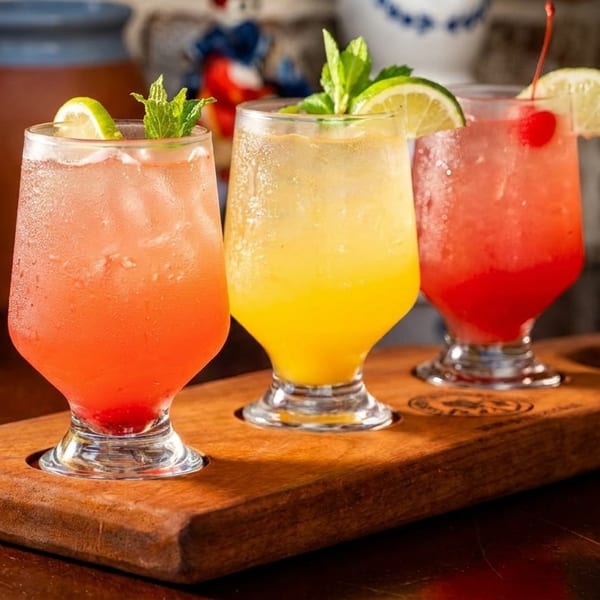 Three colorful cocktails, garnished with lime, mint, and a cherry, on a wooden serving board.