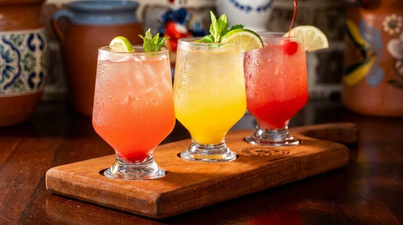 Three colorful cocktails, garnished with lime, mint, and a cherry, on a wooden serving board.