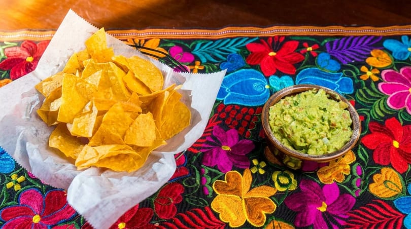 A basket of tortilla chips and a bowl of guacamole on a colorful floral tablecloth.