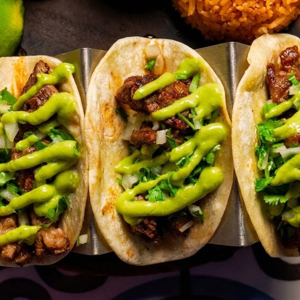 Three street tacos with meat, onions, cilantro, and green sauce, served with lime and rice.