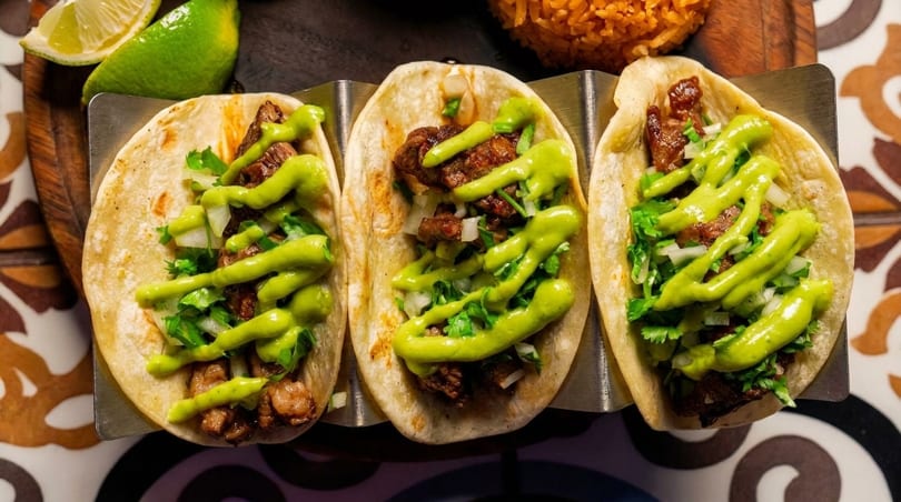 Three street tacos with meat, onions, cilantro, and green sauce, served with lime and rice.
