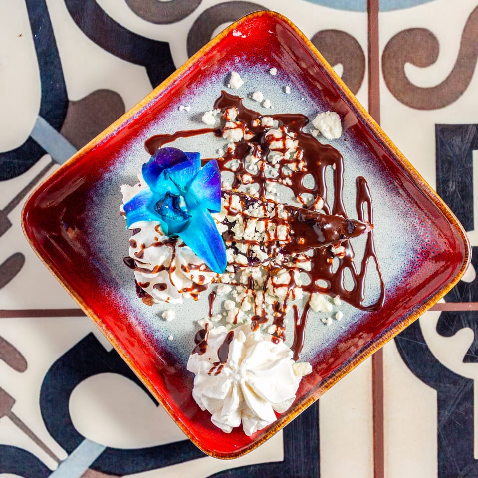 Dessert topped with whipped cream, blue edible flowers, crumbled cheese, and chocolate drizzle on a colorful ceramic plate.