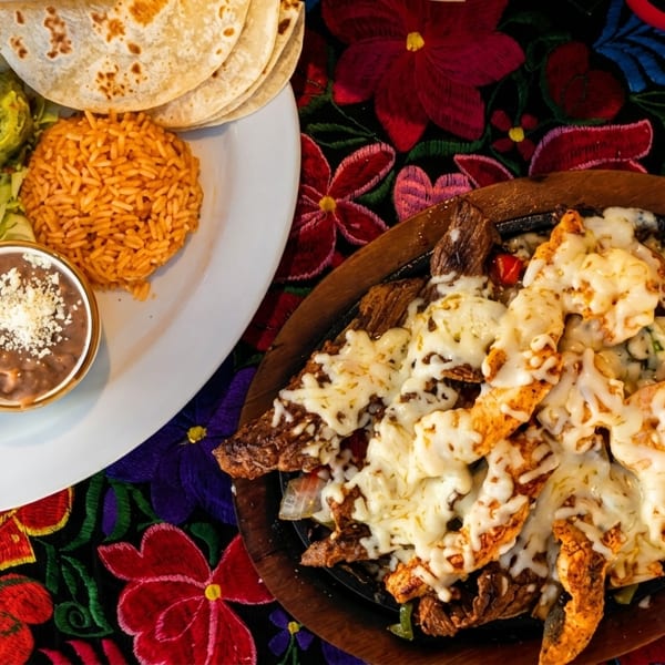 Mexican platter with rice, beans, guacamole, and fajitas with melted cheese on a colorful tablecloth.