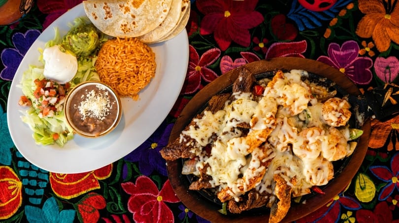 Mexican platter with rice, beans, guacamole, and fajitas with melted cheese on a colorful tablecloth.