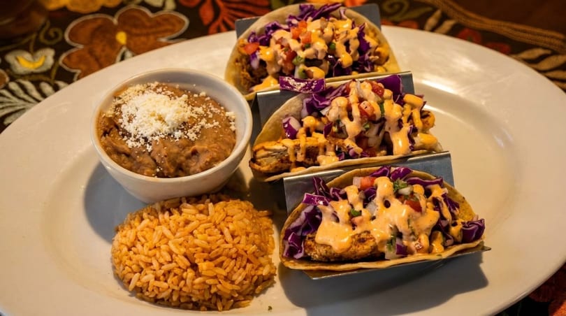 Three tacos with purple cabbage and sauce, served with rice and refried beans on a white plate.