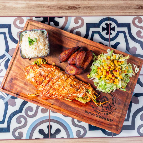 Grilled chicken platter with rice, sausage, fried plantains, noodles, and corn salad on a wooden serving board.