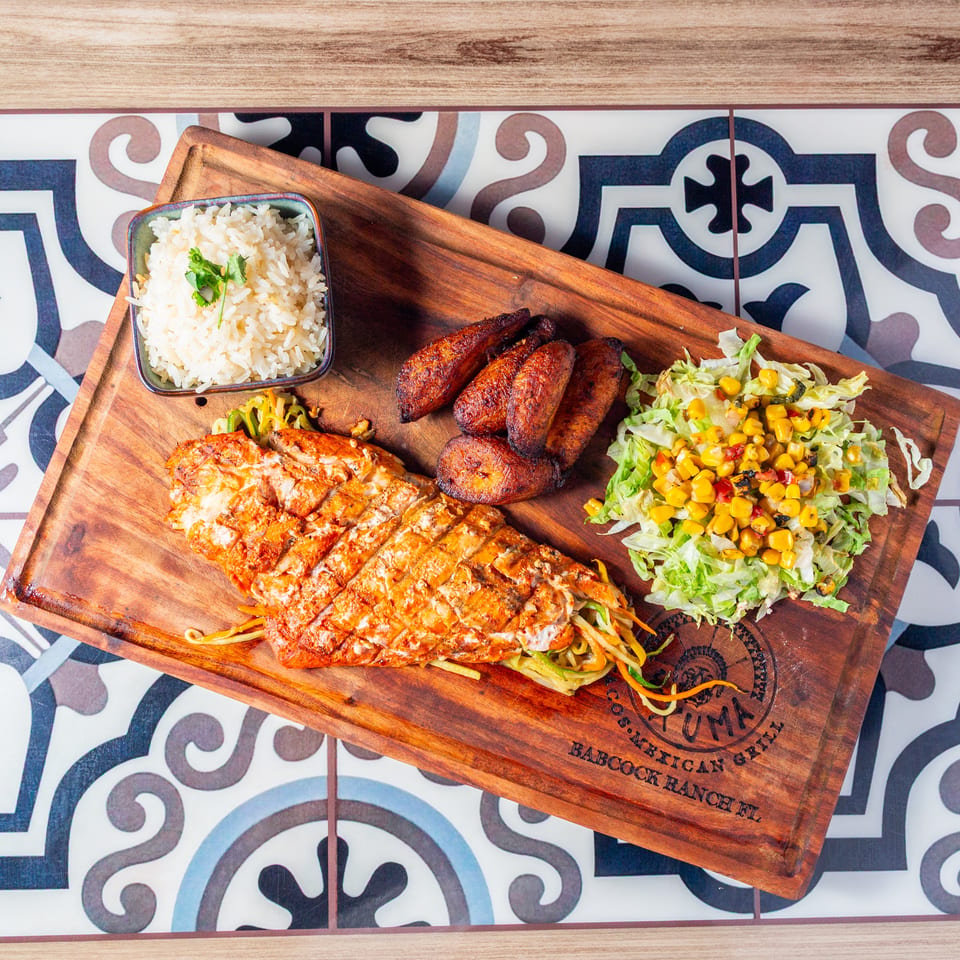 Grilled chicken platter with rice, sausage, fried plantains, noodles, and corn salad on a wooden serving board.